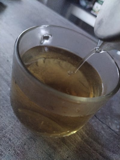 A close-up, eye-level shot shows a clear glass mug filled with a light brown liquid, likely tea, with a stream of liquid being poured into it from an unseen source, possibly a kettle. The mug sits on a textured, grey surface that resembles a wooden tabletop. The background is blurred but suggests an indoor setting with shelving and some objects, possibly in a kitchen or dining area. The lighting is dim, creating shadows and a slightly grainy texture throughout the image. The location is identified as Геранёнскі сельскі Савет, Belarus.