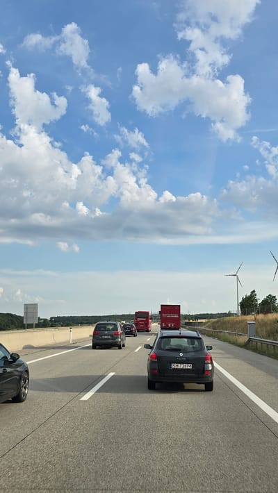 The image depicts a highway scene with multiple vehicles on a bright, partly cloudy day. In the foreground, a dark gray car is visible on the right side of the frame, facing away from the camera. To its left, a dark gray hatchback with a license plate reading "SH 73694" is also moving forward. Further ahead, another dark gray station wagon is in the left lane. Several red trucks and buses are also on the highway, including one with the text "Quehenberger" and another with "ROZVAL." In the distance, wind turbines are visible on the right side of the road. The environment appears to be a rural or suburban highway, with grassy verges and some trees. The sky is a clear blue with scattered white clouds. The license plate on the dark gray hatchback suggests the location is likely in Austria, given the "SH" prefix which is common for the state of Upper Austria. Given the context provided, the location is identified as Mistelbach, Austria.