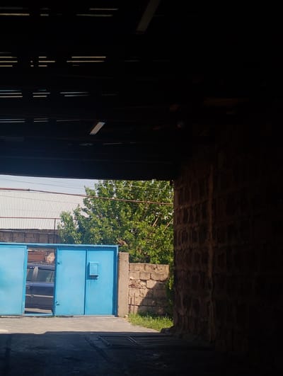 The image depicts an outdoor scene, possibly in Nor Geghi, Armenia, showing a large blue gate with a mailbox attached. To the left of the gate, a dark-colored car is partially visible. Beyond the gate, a corrugated metal roof structure and a lush green tree are observable. The foreground is shadowed, suggesting the viewer is under an overhang or inside a covered area. A rough stone wall is prominent on the right side of the image, contrasting with the smooth metal of the gate and roof. The overall impression is one of an industrial or utilitarian area, perhaps a garage or storage facility.