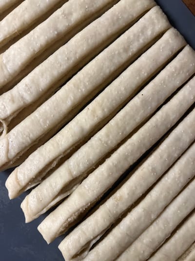 The image displays a close-up of approximately eleven parallel, elongated strips of unbaked dough. These light beige strips are arranged diagonally from the upper left to the lower right on a dark gray, rectangular baking tray. The dough surfaces are textured with fine granular particles and contain sparse minute dark specks. Each strip exhibits a layered or folded structure. The scene is located in the city of Schmittweiler, Germany. No individuals, actions, or interactions are visible. The specific indoor environment within Schmittweiler is not discernible.
