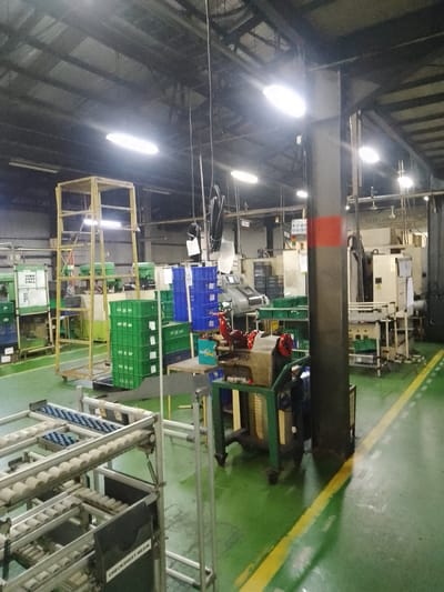 The image depicts an interior view of a factory floor in Ciantra, Indonesia. The space is industrial, with a high ceiling featuring exposed metal beams and fluorescent lighting. The floor is a vibrant green, marked with yellow and white directional lines. In the foreground, a multi-tiered conveyor system with rollers is visible. Adjacent to it is a black sign with white text that reads "CHECK SHEET MESIN."

Further into the factory, various industrial machines and equipment are present. To the left, a large machine with "POLYCON" printed on it is visible. To the right, a cream-colored industrial machine stands prominently. Stacks of green and blue plastic crates are arranged throughout the space, suggesting storage or transport of materials. A yellow metal scaffolding structure is positioned on the left side of the image. Several cables hang from the ceiling. The environment appears clean and organized, typical of a manufacturing facility. There are no people visible in the frame.