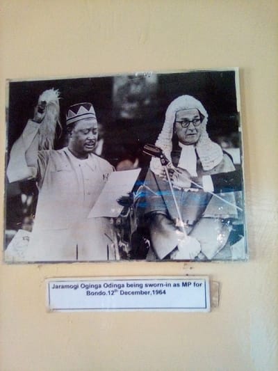 A black and white photograph, mounted on a plain, light-colored wall, depicts a historical swearing-in ceremony. The photograph itself shows signs of surface wear and reflections.
Two adult males are the primary subjects. On the left, Jaramogi Oginga Odinga, identified by the accompanying caption, is depicted mid-action. He wears a light-colored, long-sleeved tunic and a dark cap with a light-colored geometric pattern. His left hand holds a sheet of paper from which he appears to be reading, while his right arm is raised, holding a light-colored ceremonial whisk or fly-whisk. His gaze is directed downwards towards the document.
To the right, another adult male, possibly a judicial or parliamentary official, stands or sits behind a microphone on a stand. He wears a dark robe, a traditional white judicial wig, and eyeglasses. His hands are positioned in front of him, possibly resting on a desk or holding an unseen object. He appears to be observing Odinga.
The background of the black and white photograph is dark and indistinct, suggesting an indoor, formal setting. Below the main photograph, a white rectangular label with black text reads: "Jaramogi Oginga Odinga being sworn-in as MP for Bondo. 12th December, 1964."
The event depicted is Jaramogi Oginga Odinga's swearing-in as Member of Parliament for Bondo, which encompasses South Sakwa ward, Kenya, on December 12, 1964. The current image captures this historical photograph displayed on a wall, indicating its presentation as a historical record within a general area associated with South Sakwa ward in Kenya.