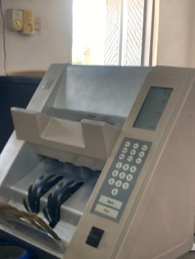 This image shows a close-up of a light-colored, upright electronic device with a digital display and a keypad. The device appears to be a piece of commercial equipment, possibly a cash counter or a ticket dispenser. The digital display shows some characters, but they are not clearly legible. The keypad features a standard number pad (0-9) and buttons labeled "Start" and "Stop".
The device is positioned in what appears to be an indoor setting, with a window or opening to the left allowing natural light to enter. In the background, a glimpse of what could be blinds or a textured wall is visible. On the wall below the window, a yellow electrical outlet with a switch is mounted.
Inside the device, a tray or compartment is visible, containing what look like dark-colored, possibly plastic, slotted objects arranged in a fanned pattern. A straw-like object is also visible next to them. The lighting suggests it might be daytime. The overall impression is of a functional space, likely a business or service area. There is no indication of people or any specific event taking place in the frame. The location context provided is Jere North, Nigeria.