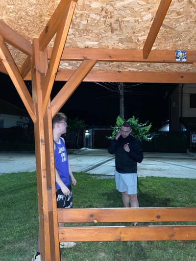 The image shows two young men standing under a wooden canopy at night. The man on the left, wearing a blue basketball jersey and patterned shorts, is partially obscured by a wooden support beam. He is looking to his right. The man on the right, wearing a black jacket and light-colored shorts, is facing the camera and appears to be adjusting something around his neck. He has his fists raised slightly. Behind them, a dark, tree-like shrub is visible, and beyond that, houses and streetlights are dimly illuminated against the night sky. The location appears to be outdoors in a residential area, possibly a yard or a public space. The ground is covered in grass, with a paved area or pathway behind it. The overall impression is of a casual, nighttime gathering.