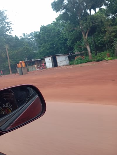 The image depicts a roadside scene in Imilike, Nigeria, characterized by a dirt road and lush vegetation. In the foreground, a car's side mirror reflects a person wearing a dark t-shirt. The mirror has the warning text "OBJECTS IN MIRROR ARE CLOSER THAN THEY APPEAR" inscribed on it. Beyond the road, a small cluster of makeshift structures, possibly shops or homes, with corrugated metal roofs are visible. These structures appear to be constructed from various materials, including wood and what seems to be tarpaulin. In front of these buildings, several large, stacked containers, likely for water or other goods, are present. A red motorcycle is parked near one of the structures. A person in a red shirt can be seen standing on the left side of the road. The environment is dominated by green trees and foliage, indicating a tropical or sub-tropical climate. The sky is overcast. The overall impression is one of a rural or semi-rural setting along a well-traveled dirt road.