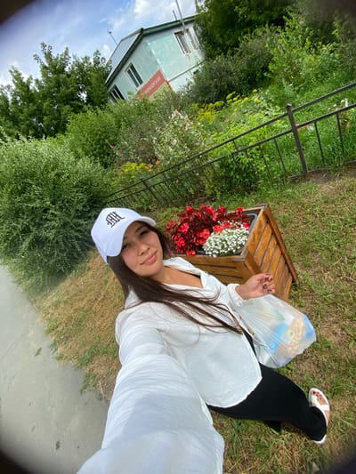 A woman wearing a white baseball cap and a white blouse is taking a selfie outdoors. She is holding a plastic bag in her right hand. Behind her, there is a wooden planter box filled with red and white flowers, a metal fence, and lush green vegetation. In the background, a light blue building with a green roof is visible, with a sign on its side that appears to be in Russian text. The woman is standing on grass next to a paved walkway. The scene is set in a natural or park-like environment within the city of Akyar, Russia.
