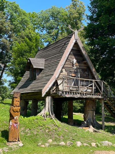 The image depicts a rustic wooden stilt house set in a lush green outdoor environment. The house has a steep, shingled roof with decorative wooden trim. A small dormer window with shutters juts out from the roof. A wooden balcony with a railing extends from the front of the house, accessible by a wooden staircase on the right. Various wooden beams and supports are visible beneath the house, some of which appear to be integrated with tree trunks. A wooden sculpture of a female figure, adorned with colorful ribbons, stands in the foreground to the left of the house. A sign with Cyrillic text hangs on the front door of the house. The surroundings are characterized by green grass, scattered rocks, and mature trees with dense foliage. The sky is clear and blue. The image appears to be taken on a sunny day. The location is identified as Litvyenski Sel'skiy Sovet, Belarus.