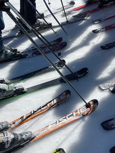 The image displays an outdoor scene at a ski resort in Jahorina, Bosnia and Herzegovina, characterized by multiple skis, ski poles, and partial views of individuals on a snow-covered surface. In the immediate foreground, two pairs of Fischer branded skis, featuring orange, white, and black graphics, are prominent, with their bindings partially obscured by accumulated snow and ice. Above these, a pair of black skis with green edge accents is attached to a dark ski boot, and further back, skis with red, white, and black patterns are connected to another dark ski boot. Multiple ski poles, colored black, blue, and silver, are visible throughout the frame; some are held upright by unseen individuals, while others stand in the snow, displaying yellow or lime green baskets. The lower extremities of at least two individuals are present in the upper left quadrant, clad in dark and blue ski pants with dark ski boots. The packed snow surface is illuminated by strong sunlight, casting distinct diagonal shadows from the people and their equipment.