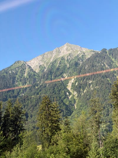 This is a vertical, eye-level, medium shot of a mountain landscape in Scharnachtal, Switzerland. The image is dominated by a steep, rocky mountain face on the left, covered in sparse vegetation and scree slopes. To the right of the mountain, the terrain slopes down and is densely covered with a thick forest of coniferous trees. The sky is a clear, bright blue, indicating a sunny day. A rainbow-like lens flare streaks diagonally across the mountain and forest, casting a reddish hue. The lighting suggests it is daytime, likely mid-morning or afternoon, given the intensity of the sunlight. The scene evokes a sense of natural grandeur and the rugged beauty of the Swiss Alps. No people, objects, or text are visible in the image.