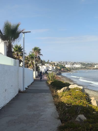 This image depicts a paved walkway alongside a white stucco wall and lush vegetation, leading towards a beach. A person is seen cycling on the walkway, heading away from the camera. In the background, palm trees and white buildings are visible, with a coastal town or city nestled on a hillside overlooking the sea. The ocean has small waves gently rolling onto a pebble beach. The sky is clear and blue, suggesting a pleasant day. The sun appears to be shining, casting shadows and illuminating the scene, indicating it's likely morning or late afternoon. The overall atmosphere is serene and vacation-like, consistent with a coastal resort in La Gaspara, Spain. No visible text is present in the image.