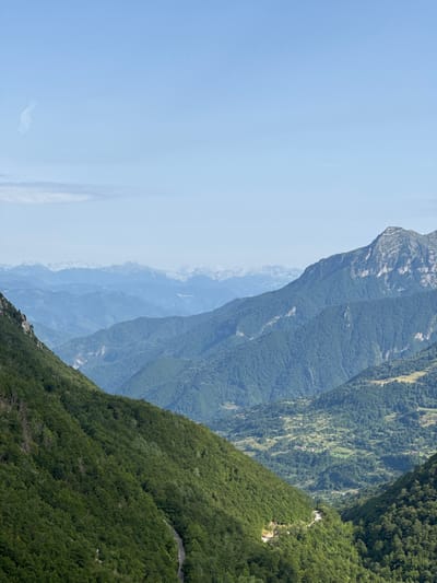 The image depicts a vast, mountainous landscape under a clear blue sky with a few wispy clouds. The foreground is dominated by a steep, densely forested mountainside on the left, with a winding road visible. The middle ground features rolling, tree-covered hills and a valley with scattered houses and agricultural fields. In the background, a series of progressively lighter blue mountain ranges recede into the distance, suggesting a significant elevation and a hazy atmosphere. The scene is devoid of people or specific man-made structures, emphasizing the natural, rugged environment. The lighting suggests it is daytime, likely mid-morning or afternoon, with the sun casting light and shadows that highlight the contours of the terrain. The overall impression is one of natural grandeur and a remote, untouched wilderness.