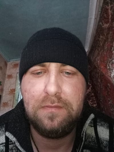 A close-up, eye-level, indoor selfie of a Caucasian male wearing a black knit beanie. He has a short, unkempt beard and light-colored eyes. He is wearing a dark-colored sweater with a light-colored pattern. The background consists of a wall with floral wallpaper on the left and a patterned rug or tapestry on the right. The location is potentially Parabel, Russia, based on the user's prompt. The ceiling is painted a pale blue or green color. The subject appears to be looking down and appears to be slightly sad.