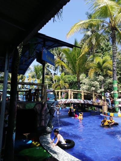 The image depicts a lively scene at a water park, likely located in Tegalwangi, Indonesia, suggested by the lush tropical vegetation and palm trees. In the foreground, a young boy in a white tank top and dark shorts is sitting on the edge of a bright blue pool, reaching towards a black inner tube in the water. The pool is filled with people, many of whom are children wearing colorful swimwear and floatation devices, suggesting active recreation.
To the right, two young boys in yellow shirts are sitting on black inner tubes, seemingly enjoying the water. In the middle ground, a wooden bridge with a decorative stone railing spans the pool. Several individuals, including adults and children, are visible on and around the bridge. Further in the background, a water slide structure with blue elements can be seen.
Above the pool area, a covered structure with a corrugated roof partially shades the scene. A blue sign with white text that reads "AYO KARAOKE" is prominently displayed. The overall environment is one of a recreational facility with a vibrant, sunny atmosphere. The water is a clear, inviting blue, and the surrounding greenery provides a natural, tropical backdrop. The presence of a karaoke sign hints at the entertainment offerings of the water park.