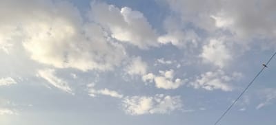 The image displays a vast expanse of sky filled with scattered white and gray clouds, suggesting it could be a partly cloudy day. The lighting indicates it might be late afternoon or early morning, with the sun illuminating some of the clouds from behind. In the lower right quadrant of the image, a thin black wire stretches diagonally across the frame. Perched on this wire is a small bird, facing towards the left. The bird appears to be dark with some lighter markings on its chest and head. The background is a soft blue sky, with a wider, more diffused cloud formation filling most of the upper portion of the image. The location is identified as Margilan City, Uzbekistan.