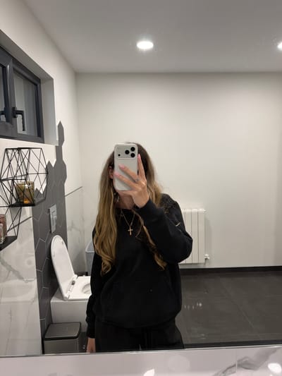 A woman is taking a mirror selfie in a bathroom. The bathroom has white walls, grey tiled flooring, a radiator, a toilet with the lid up, a trash can, and a window. The woman is wearing a black hoodie and a gold necklace with a cross. She is holding a white iPhone in front of her face. The location appears to be a modern bathroom, but there is no indication of being in the city of Resilovo, Bulgaria.
