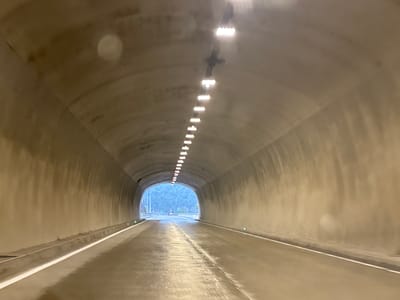The image shows the interior of a tunnel on a road, likely in or near Grigalati, Georgia. The tunnel is constructed of concrete and is illuminated by a series of bright lights running along its ceiling. The road surface appears to be wet, reflecting the lights, and there are white lines marking the lanes. The tunnel extends into the distance, with a bright, natural light source visible at the end, suggesting the exit of the tunnel. The walls of the tunnel are textured concrete, and there are small green lights embedded in the sides of the road near the exit. The scene is captured from the perspective of someone traveling through the tunnel, likely in a vehicle. There are no people or other vehicles clearly visible within the tunnel. The environment outside the tunnel appears to be mountainous, with trees visible.