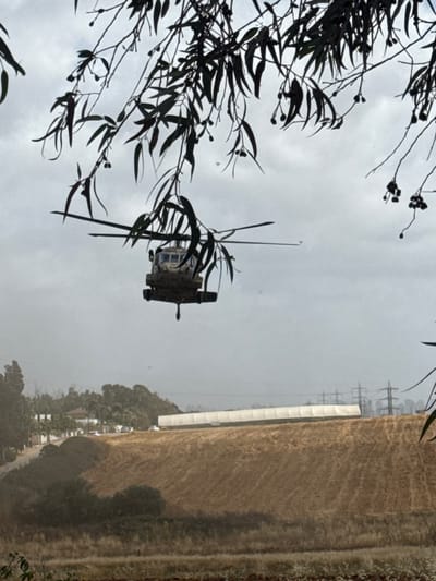 A blackhawk helicopter is hovering in the air, seen from below. It is flying over a large, flat, dry, brown field. The field is surrounded by trees and a white building in the distance. Power lines are visible in the background.  The photo was taken from a leafy tree branch in Ramot HaShavim, Israel, at the location specified by the coordinates 32.162, 34.874. The sky is overcast with gray clouds.  The helicopter is the primary subject of the photo.  The trees, power lines, and field are background elements. The helicopter is stationary and the only visible object in motion.  There are no people visible in the image.  The scene is calm and peaceful.