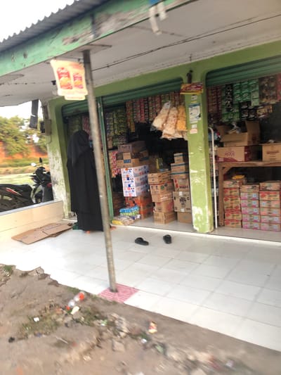 A full-length view shows a street-level storefront in Sentul, Indonesia, with the green painted facade of a shop. A woman wearing a black niqab and abaya stands at the entrance to the shop. The shop is filled with merchandise, including boxes of food and beverages stacked on shelves and on the floor. The exterior of the shop has a covered awning and a pole that supports a sign with images of food. The ground in front of the shop is tiled and there are some cardboard boxes and a plastic bottle on the ground to the left. A motorcycle is parked on the left side of the image, near the storefront. The street in front of the shop appears to be paved with white tiles. The scene is likely a small convenience store or warung.