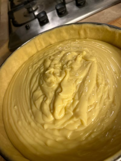 A close-up shot reveals a pie crust filled with a creamy, light yellow custard. The custard has a glossy sheen and is marked with subtle lines where it was poured or spread. Dark flecks, likely vanilla bean seeds, are visible throughout the custard. The crust is a pale, slightly rough texture, contrasting with the smooth filling. A portion of a metallic object, possibly part of a stovetop, is visible in the lower left corner.