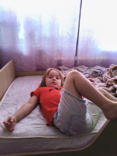 A young child with light brown hair, wearing a red t-shirt and light blue denim shorts, is lying on their back on a mattress. The child is looking towards the camera with a neutral expression. The mattress appears to be on a bed frame. In the background, there are rumpled blankets and sheets, and a curtain or fabric hanging vertically behind the bed. The lighting suggests it could be daytime, possibly indoors with natural light coming from behind the curtain. The overall scene is casual and domestic. There is no visible text in the image.