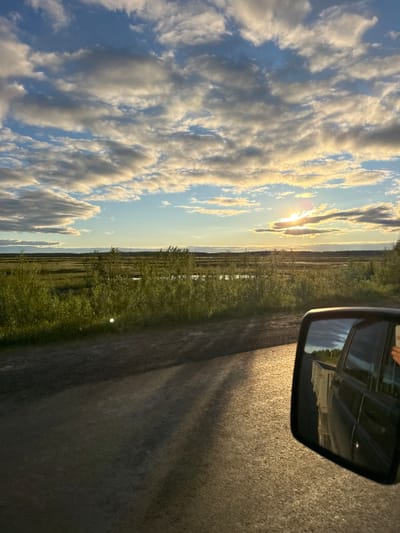 The image depicts a view from inside a vehicle, looking out at a rural landscape during what appears to be sunset or sunrise. The camera is oriented vertically, suggesting the photo was taken with the phone rotated.

The main subjects include the sky, filled with dramatic, golden-tinged clouds against a blue background, suggesting a dramatic weather event or the golden hour. Below the sky, a flat, green landscape stretches out, dotted with scattered trees or bushes. A body of water, possibly a pond or marsh, is visible in the distance. To the right of the landscape, a portion of the vehicle's exterior is visible, including a side mirror reflecting the sky and the interior of the car. The road surface, which appears to be asphalt or gravel, takes up the lower right portion of the image, showing dappled sunlight and shadows. The overall mood is serene and reflective, capturing the beauty of nature during a transitional time of day. The location context of Parma, Russia, implies a rural or wilderness setting. There is no visible text in the image.