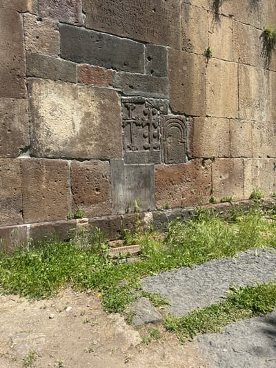 The image shows a stone wall with carvings and inscriptions, located at the St. Gregory Church in Bjni, Armenia. The wall is constructed from large, rough-hewn stones of varying colors, including shades of brown, grey, and a reddish-brown. There are multiple carved elements on the wall, including what appear to be crosses and other geometric or symbolic designs. Inscriptions in an ancient script are visible on some of the upper sections of the wall. The lower portion of the wall is partially overgrown with green grass and other vegetation. The ground in front of the wall is a mix of dirt and some stone paving. Sunlight casts shadows on the wall, suggesting it is daytime. There are no people or animals visible in the image.