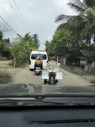 In Teupinreudeup, Indonesia, the image captures a scene on a dirt road with a white van and two motorcycles. The van, a Toyota Hiace, is parked on the road, with two people on a motorcycle behind it. The driver of the motorcycle is wearing a light brown shirt and a grey cap, while the passenger wears a yellow shirt and a white cap. Another motorcycle with a sidecar carrying what appears to be food items is positioned in front of the van. The sidecar has white panels with text, and the motorcycle is ridden by a person wearing a light brown shirt and a grey cap. The environment is rural, with lush green trees and vegetation lining the road. The sky is overcast. The photograph is taken from inside a vehicle, showing the dashboard and windshield.