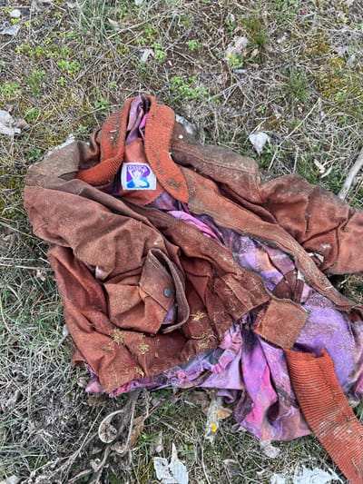 A heavily soiled, crumpled, reddish-brown suede-like jacket with a rust-colored ribbed knit collar and cuffs lies discarded on sparse, dry grassy ground mixed with dirt, twigs, and plant debris in Stoianovo, Bulgaria. The jacket is unzipped, revealing a vibrantly patterned purple garment with pink and orange abstract designs underneath. A blue brand label affixed to the interior collar of the jacket displays the white text "YESSICA" above a white silhouette of a woman's head. The jacket's exterior shows substantial dirt and plant matter accumulation, particularly on the ribbed collar and lower sections. A single flap pocket with a dark, circular snap button is visible on the jacket's lower left side. One rust-colored ribbed cuff is visible detached or torn from the jacket at the bottom right. No individuals are present.