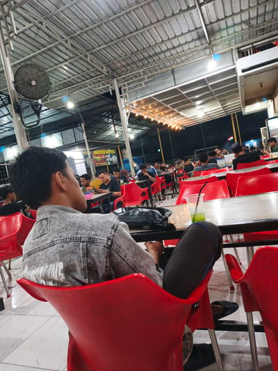 This image depicts a lively outdoor dining area, likely a food court or restaurant in Cotpuuk, Indonesia, during the evening. The setting is characterized by a large, corrugated metal roof supported by metal beams, providing shelter from the elements. Rows of bright red plastic chairs surround tables, many of which are occupied by people.
The primary subjects are the patrons of the establishment. In the foreground, a young man is seen from the back, wearing a denim jacket and dark pants, seated in a red chair. His gaze is directed away from the camera, suggesting he might be looking at something or someone else. Other individuals are scattered throughout the scene, some engaged in conversation, others looking at their phones, and a few appear to be eating. The atmosphere seems casual and social, indicating a typical evening gathering.
A large industrial fan is mounted on a pole on the left side of the frame, suggesting a warm climate where ventilation is important. Overhead, strings of warm, yellow lights are strung across the ceiling, illuminating the area and creating a festive ambiance. In the background, there are signs and structures that hint at a commercial area. One sign, partially visible, displays Indonesian text.
The time of day is clearly evening or night, given the artificial lighting and the dark sky visible in some areas. The weather appears to be fair, as people are dining outdoors. The overall impression is one of a busy and popular spot for people to gather and enjoy a meal or drinks in a relaxed, communal setting. The presence of numerous tables and chairs, along with people actively dining, suggests an active food establishment.