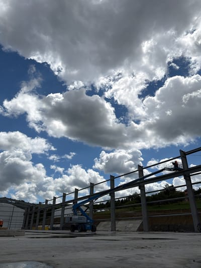 In Skrzydlna, Poland, a construction site is captured under a dynamic sky filled with dramatic white and gray clouds against a vibrant blue backdrop. The foreground consists of a wet, unfinished concrete surface. A large, blue Genie boom lift is positioned centrally, its arm extended upwards towards the steel framework of a building under construction. A construction worker, wearing an orange vest, is visible in the basket of the boom lift, actively working on the structure. The unfinished building features prominent steel beams and vertical supports. In the background, a gentle slope of green grass is visible, dotted with trees and a distant house. Another construction worker in an orange vest is standing near the lower left corner of the frame, by a concrete barrier.