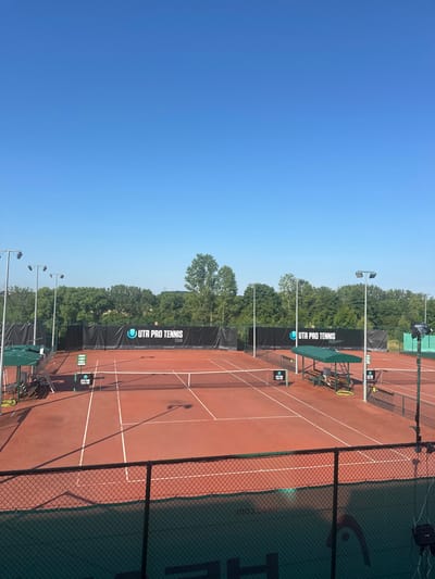 The image displays an outdoor professional tennis facility located in Giebułtów, Poland. The scene, captured under a clear, bright blue sky, features at least three red clay tennis courts, meticulously marked with white lines. Each court is equipped with a dark tennis net.
Prominently displayed along the back boundaries of the courts are black banners featuring the white "UTR PRO TENNIS TOUR" logo, indicating a professional-level competition venue. Behind these banners, a dense line of green trees forms the background. Numerous tall light poles, each fitted with multiple luminaires, are positioned around and between the courts, suggesting the facility is equipped for night operations.
On the left side of the courts, a green market umbrella and associated seating are visible. On the right, a more substantial green-roofed structure with benches provides shaded seating. A coiled green water hose is present near this structure. Small, vertical digital displays or scoreboards, also bearing the "UTR PRO TENNIS" logo, are positioned near the nets on multiple courts. In the lower foreground, a black chain-link fence is partially covered by a dark green mesh windbreak or banner displaying a white "HEAD" logo. In the lower right corner, a portion of professional camera or broadcasting equipment is visible behind this foreground fence. No individuals are present on the courts or within the immediate viewing area. The facility appears prepared for use but is currently unoccupied. This setup is consistent with the infrastructure of a professional tennis club such as KT Błonia Racquet Park, known to host UTR events in the region.