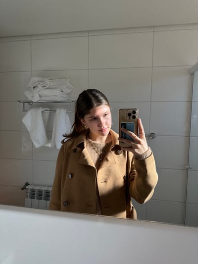 A female subject is depicted in a bathroom, identified as being located in the city of Canela, Brazil, taking a mirror selfie. The subject, visible from the chest up, has light brown hair and is wearing a tan double-breasted coat over a light brown knitted sweater. She accessorizes with gold hoop earrings, a pendant necklace, and multiple rings on her right hand. She holds a gold-colored smartphone with a clear case in her right hand, with a blue card visible within the case. The background consists of white tiled walls with a chrome towel rack on the left holding folded and draped white towels. Below the towel rack, a white heating radiator is visible. The foreground features the white reflective surface of a bathtub rim or counter, with part of a glass shower enclosure visible on the far right. Natural light from the right casts shadows across the subject and the tiled wall.