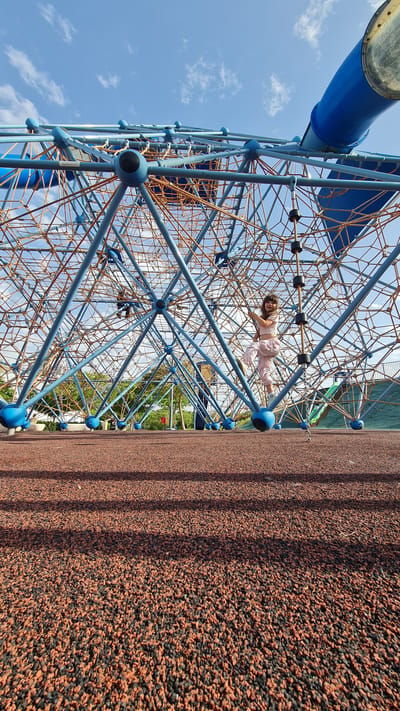 A wide-angle, rotated, upward view captures a child on a playground climbing structure, presumably in Kiryat Gat, Israel. The child, wearing light pink clothes, is positioned within the blue metal frame and orange rope net of the climbing structure. The background includes a blue sky with scattered clouds. The playground surface on the right side of the frame is a dark reddish-brown material. The image is angled to show the height of the structure, and the child appears to be suspended in mid-climb. A blue tube, possibly a slide entrance, is visible in the top left corner. Green foliage suggests the playground is near trees or landscaping.