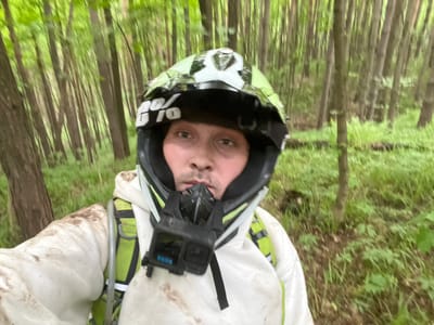 A man in a dirt bike helmet and white protective suit stands in a forest. The helmet has a GoPro attached to the top. The background shows closely packed trees and green vegetation, suggesting a wooded area. The man is facing the camera directly. The trees resemble the kind of forest that grows in Russia. The location of Gruzinskii, Russia, cannot be determined from the image.