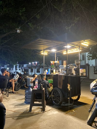 The image depicts a nighttime scene at a mobile coffee stand located on a street in Gampongjawa, Indonesia. The coffee stand is illuminated by bright overhead fluorescent lights, casting a warm glow on the equipment and the surrounding area. The stand itself is a dark, wheeled cart with a silver awning. On the counter, there are multiple coffee machines, including an espresso machine and a grinder filled with coffee beans, along with various bottles and containers. A large blue water cooler and a brown plastic stool are positioned to the left of the stand.
Several individuals are present. Three men are seated on chairs to the left, facing away from the camera and towards the coffee stand, appearing to be customers. One man, wearing a black shirt, is seated at the stand, seemingly engaged in preparing drinks. Another man, also in a black shirt, is visible from behind on the right side of the stand. Motorcycles are parked to the left of the coffee stand and a portion of a motorcycle is visible in the lower right corner of the image. The ground is paved concrete, with scattered leaves. Trees with visible branches and foliage overhang the scene. In the background, there are buildings with lights on, suggesting a residential or commercial area. The overall atmosphere is casual and social, typical of a street-side eatery at night.