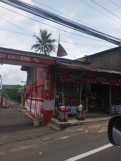 This image, located in the city of Chori, Nigeria, presents a street-level view of a commercial area under a partly cloudy sky. The scene is dominated by a two-story structure, primarily painted red and white, with a corrugated metal roof showing signs of rust. An archway extends from the left side of this building, displaying the text "CO RANJENG 01/05" in white lettering on a red background. Above the shop section, a flagpole displays a red and white horizontal bicolor flag.
The building's ground floor appears to be a shop or kiosk, secured by vertical metal bars. Multiple banners advertising "PRIMA" and "BARU" with a price of "15.000" are strung across the shop's facade. Various packaged goods are visible inside.
A single individual, wearing dark clothing and a head covering, sits on a concrete ledge outside the shop. They are positioned behind two red-framed portable stalls. One stall holds a large transparent water dispenser bottle, and the other contains a metal steaming pot. Two white plastic buckets rest on the ground near the building's corner. The building's side wall features painted numbers "17," "08," and "45" in black on a white background.
To the left of the building, a paved road slopes downward, revealing a series of descending terraces or steps with red and white railings, leading into an area densely covered with green trees. A tall palm tree is visible in the background, towering above the buildings. The foreground shows a section of asphalt road with a white demarcation line, and the edge of a dark vehicle's rearview mirror is visible on the far right, suggesting the photograph was taken from within a vehicle. Overhead, numerous electrical or utility wires crisscross against the sky.
