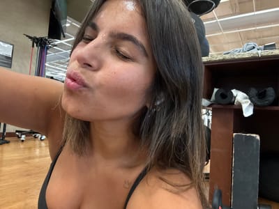A female individual is depicted in a selfie, presumably taken within a fitness facility in Plainsboro Township, United States. Her face, showing visible perspiration on the forehead and nose, is positioned with eyes closed and lips puckered. She has long, dark brown hair and wears a black top, revealing a small crown tattoo on her upper left chest. A white earbud is present in her right ear. The background features light wood-patterned flooring. To the left, a mobile rack holds multiple colored resistance bands. To the right, a brown shelving unit contains various items including a grey garment, a white towel, and black cylindrical objects resembling foam rollers or equipment covers. Overhead lighting fixtures are partially visible in the upper background.