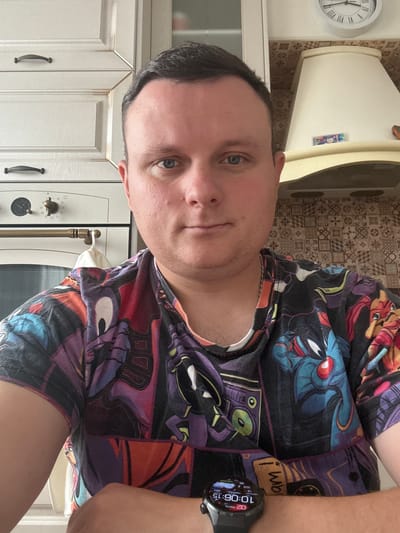 The image shows a selfie of a person in a kitchen. The person is a white male with short brown hair and blue eyes. He is wearing a t-shirt with a colorful cartoon print. He has a smartwatch on his left wrist. The background shows kitchen cabinets, a stove, and a range hood. The time on the smartwatch is 10:06. The scene is indoors, in a domestic setting. This image was taken in Kondopoga, Russia.