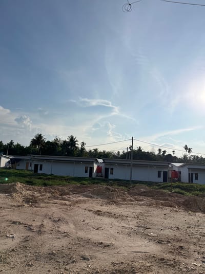 The image depicts a row of white, single-story buildings with dark-colored doors and windows, possibly residential units, under a bright blue sky with scattered clouds. In the foreground, a dirt field with tire tracks and debris suggests a construction site or undeveloped land. A few palm trees and other vegetation are visible behind the buildings. A red water tank is positioned next to one of the structures. The overall setting appears to be in a tropical or subtropical climate, consistent with the location of Alurcucur, Indonesia. There are no people or discernible activities captured in the frame.
