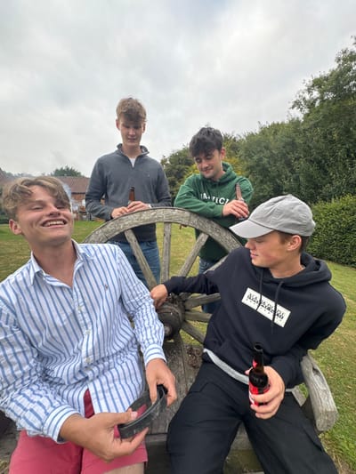 Four young males are depicted outdoors in Bishop Wilton, United Kingdom, under an overcast sky, gathered around a large, weathered wooden wagon wheel.
In the foreground on the left, a male with light brown hair, wearing a blue and white striped collared shirt and red shorts, is positioned leaning or seated, smiling with his head turned to the left. He holds a black, curved, rubber-like object in both hands, and a small, smoldering item, emitting a faint wisp of smoke, is visible between his fingers near his jaw. To his right, a male wearing a black hoodie with a white "NAPAPIJRI" logo, black cargo pants, and a light grey baseball cap, leans on the wooden wheel, holding a dark glass bottle with a red label in his right hand, and looks towards the first male.
Behind the wooden wheel, two additional males stand. The male on the left wears a grey half-zip sweater and blue jeans, holding a dark glass bottle in his right hand and looking towards the viewer. The male on the right wears a dark green hoodie with white text, holding a dark glass bottle, and is looking slightly downwards.
The ground is covered in green grass. The background features dense green foliage and a portion of a reddish-brown brick building with a white-framed window visible on the left side of the frame.