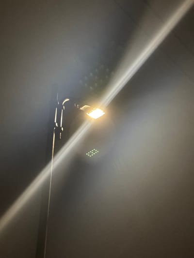 A photograph captures a bright, diamond-shaped LED light fixture illuminating a dark, indistinct environment. The light emits a strong, warm white glow, creating prominent diagonal light rays or lens flares that traverse the image. The fixture is mounted on a dark, bent metal pipe or bracket, which is secured to a horizontal bar, suggesting an industrial or utility setting such as a garage, underpass, or indoor facility in Engomi, Lefkoşa, Cyprus.

To the left and slightly below the main light, a cluster of softer, hexagonal or circular reflections is visible, likely an artifact of the light's internal structure or lens flare. More strikingly, to the right and slightly above the light, a distinct pattern of small, bright green dots appears. These dots are arranged in a sparse, rectangular grid-like formation, possibly another type of lens flare or internal reflection against the muted, dark background. The overall scene is characterized by darkness, indicating night or an enclosed space lacking natural light. A small, unreadable white label or tag is visible hanging from the light fixture. There are no discernible people or specific activities taking place, focusing solely on the artificial light source within its environment.