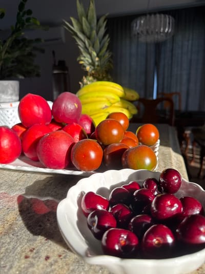 A still life composition features a variety of fruits arranged on a granite countertop, bathed in direct sunlight. In the foreground, a white, fluted bowl overflows with dark red cherries. Behind this bowl, a white rectangular plate holds a collection of red fruits, possibly plums or peaches, interspersed with a few dark red, round fruits resembling cherry tomatoes. Further back, a bunch of yellow bananas and the leafy crown of a pineapple are visible. The background is dimly lit, showing a dark curtain and furniture, suggesting an indoor setting, likely a kitchen or dining area. No people or animals are present in the image. There is no information within the image to suggest a location in Dieppe, Canada.