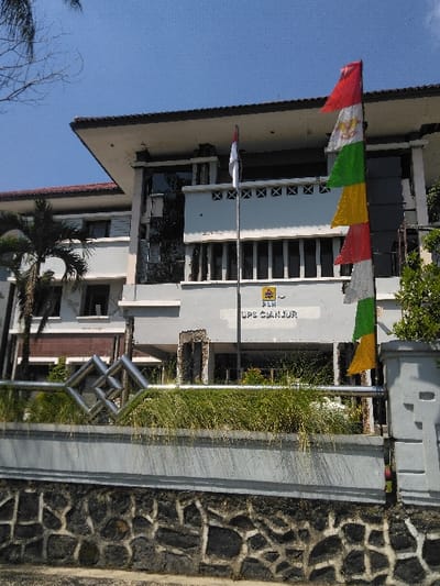 The image depicts the exterior of the PLN UPS Cianjur facility, located in Cianjur, Indonesia. No individuals are present or engaged in any discernible action or interaction. The multi-story building features a grey and white facade with a brown roof and numerous windows, some reflective. The central ground-level entrance displays exposed concrete and rebar, indicative of damage or ongoing construction. Two flagpoles stand in front of the building; the left pole displays the red and white flag of Indonesia, while the right pole holds a vertical series of small rectangular pennants in red, white (with an emblem), green, yellow, red, and white. The foreground consists of a decorative metal railing with a diamond pattern, a planter box containing tall green grass, a grey concrete wall, and a dark, irregular stone retaining wall. A palm tree is visible on the left, with additional green foliage on the right. The scene is illuminated under a clear blue sky.
