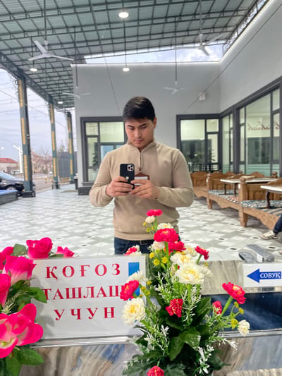 The image depicts a male individual, appearing to be in his 20s or 30s, with short dark hair, reflected in a polished surface, possibly a mirror or large glass panel, within a commercial establishment in Chinabad, Uzbekistan. The man, wearing a light tan quarter-zip sweater and blue jeans, holds a black smartphone with both hands, angled as if photographing the reflection itself. A brown lanyard is visible around his neck.
In the actual foreground, closest to the viewer, is a white rectangular sign with red Cyrillic text reading "ҚОҒОЗ ТАШЛАШ УЧУН" (FOR PAPER DISPOSAL). To the right of this sign, a blue arrow points right with the Cyrillic word "СОВУК" (COLD). Artificial floral arrangements featuring vibrant pink/red and white flowers, along with green foliage, are positioned around the sign. The surface beneath these items appears to be a light-colored, possibly marble, counter.
The reflected background reveals an open-air or semi-covered patio area transitioning into an indoor space. To the right of the reflected man, an indoor dining area is visible through large glass panels, furnished with carved wooden tables and benches with upholstered cushions. The floor of this indoor space features a grey and white geometric tiled pattern. To the left, beyond the reflected man, an outdoor street scene is visible under a cloudy sky, showing distant buildings and parked vehicles. The overhead structure is a metal framework supporting a corrugated roof, from which multiple white ceiling fans and recessed light fixtures are suspended.
