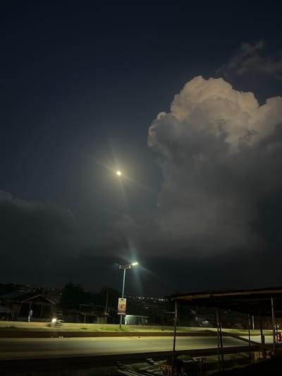 The image shows a nighttime scene in Offatedo, Nigeria. A street is visible on the right side of the frame, illuminated by a street lamp. Buildings and structures line the street. The sky is dominated by dark clouds with a bright moon.