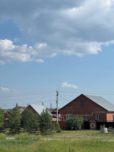 This is a vertical, outdoor, daytime photograph of a residential area in Prokudskoye, Russia, under a partly cloudy sky. The image features several houses with red brick exteriors and dark metal roofs, characteristic of Russian rural architecture. Green grass covers the ground around the houses, with some trees and bushes also visible. Power lines and utility poles are present, suggesting the presence of modern infrastructure. The overall impression is one of a peaceful, rural setting. The time of day appears to be midday, based on the bright sunlight and clear skies, though some clouds are present. There is no text visible in the image.