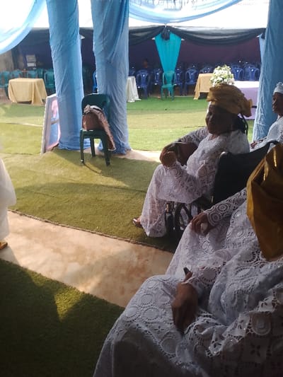 This image depicts an outdoor gathering in Ufuma, Nigeria, beneath an expansive temporary canopy structure. The ground surface is composed of artificial green turf, with a distinct concrete walkway traversing the foreground. In the midground, a woman is seated in a black wheelchair. She wears a white lace dress featuring intricate patterns and a prominent gold headwrap. Her left hand holds a small, light-colored object, while her right hand rests on her lap, holding a white object. Her gaze is directed towards the left side of the frame. Partially visible in the right foreground is another individual, similarly attired in a white lace garment and a gold headwrap, with a white object visible in their right hand.

To the left of the woman in the wheelchair, a dark green plastic chair stands, supporting a brown purse with a patterned strap. A white display board with a blue border is partially visible near this chair. The canopy structure in the immediate foreground is characterized by large columns wrapped in light blue fabric. The background reveals a more expansive tent area, draped with black and light blue fabric, under which numerous blue plastic chairs are arranged. Several tables covered with light-colored tablecloths are interspersed among the seating, one of which displays a white floral centerpiece. Additional individuals are seated in the background, observing the event.