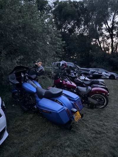 In Mensdorf, Luxembourg, a gathering of motorcycles and a car is depicted outdoors on a grassy area adjacent to a tree line. In the foreground, a bright blue motorcycle is parked, with its license plate reading "HD 1812". Behind it, two other motorcycles, one dark red and one dark maroon, are visible. A dark-colored car is parked in the background, with distinctive graphics on its side. The scene is illuminated by ambient light, suggesting it is either early morning or late evening.