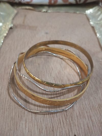 **Location:** Chandragiri, Nepal.

**Description:** A close-up, eye-level photograph depicts seven bangles resting on a light-colored wooden surface. Four bangles are gold-toned; one features an etched geometric pattern of repeating triangles, and another displays an etched floral or leaf pattern. The remaining two gold-toned bangles appear smooth. Three bangles are silver-toned, exhibiting a thinner profile and a textured, possibly hammered finish. The bangles are loosely intermingled, with some partially overlapping others. The wooden surface, possibly a tabletop or floorboard, shows visible grain patterns. The background is blurred, revealing a light-toned surface with indistinct, lighter-colored patterns, potentially fabric or paper. No individuals or actions are depicted.