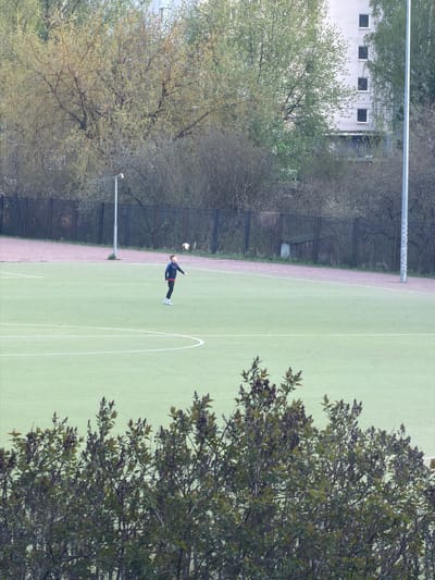 A horizontal aerial shot depicts a soccer field in Myaglovo, Russia. The field is surrounded by trees and a building is visible in the background. A person is kicking a soccer ball on the field.